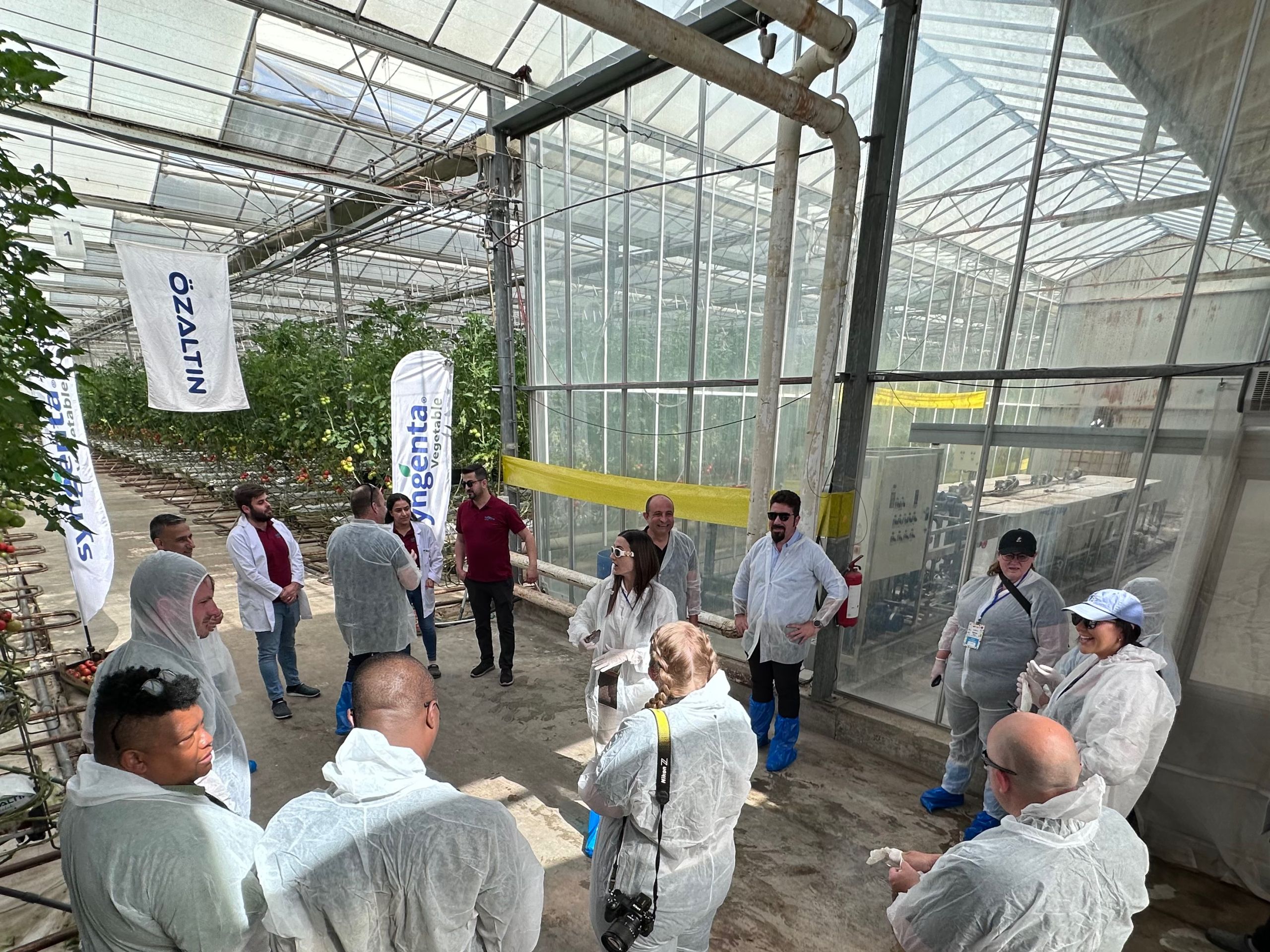 White-coated journalists listening to a presentation inside a greenhouse.
